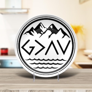 May include: White ceramic plate with a black mountain and wave design, and the text "G > AV". The plate has a black border and is displayed on a clear stand. Kitchen decor.