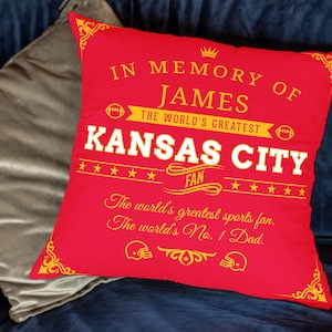 May include: Red throw pillow with gold lettering that reads "In memory of James, the world's greatest Kansas City fan. The world's greatest sports fan. The world's No. 1 Dad."
