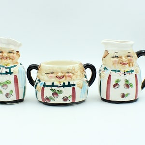 May include: A set of three ceramic sugar and creamer containers featuring whimsical cartoon faces with white hats and aprons. The containers are decorated with red, green, and blue accents and feature a fruit motif.
