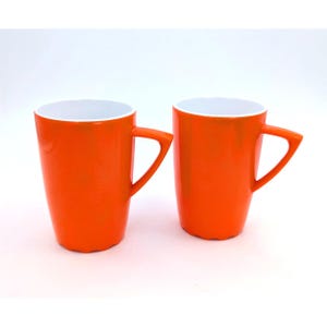 May include: Two bright orange mugs with a smooth, glossy finish and a white interior. Each mug features a distinctively angular, triangular-shaped handle and a slightly scalloped base, standing side by side against a plain white background.