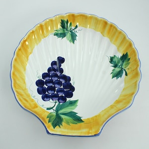 May include: A white ceramic serving dish shaped like a seashell with a yellow rim and a blue and green grapevine design. The dish features a cluster of dark blue grapes.