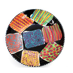 Könnte beinhalten: Ein schwarzer Keramikteller mit einem weiß-schwarz gestreiften Rand. Der Teller zeigt sieben farbenfrohe, rechteckige Designs mit verschiedenen Mustern, darunter Streifen, Karos und Punkte. Die Designs verwenden Farben wie Rot, Gelb, Grün und Lila.