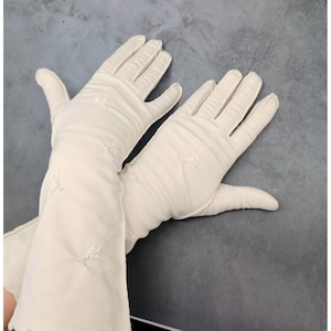 Könnte beinhalten: Ein Paar ellenbogenlange Handschuhe in Cremeweiß mit zarten, kleinen weißen Blumenstickereien, die über den Stoff verteilt sind. Die Handschuhe haben eine glatte, matte Oberfläche und reichen über das Handgelenk bis zum Unterarm.