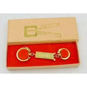 Könnte beinhalten: Goldfarbener Schlüsselanhänger mit zwei Schlüsselringen und einem rechteckigen Anhänger mit der Aufschrift "J.L.B.". Der Schlüsselanhänger befindet sich in einer rot-weißen Schachtel mit der Aufschrift "Custom Crafted" auf dem Deckel.