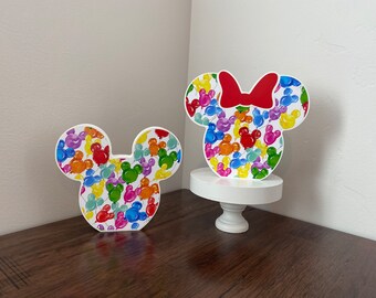 May include: Two white cardboard Mickey Mouse and Minnie Mouse head cutouts with a colorful balloon pattern. Minnie Mouse has a red bow.