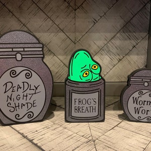 May include: Three decorative jars with glitter and black paint. The jars are labeled "Deadly Nightshade", "Frog's Breath", and "Worm's Wort".