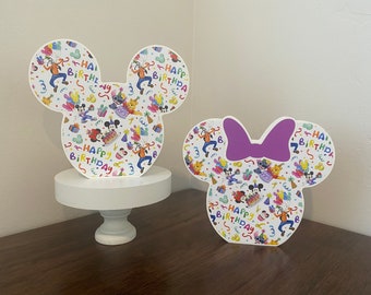 May include: Two white cardboard Mickey Mouse and Minnie Mouse head cutouts with a colorful pattern of Disney characters and the text "Happy Birthday".