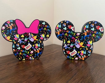 May include: Two black Mickey Mouse head shaped decorations with a colorful birthday theme. One decoration has a pink bow on top. Both decorations have the text "Happy Birthday" on them.