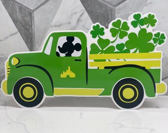 May include: A green truck with yellow detailing, carrying a load of green shamrocks. The driver's side window features a silhouette of Mickey Mouse. A yellow castle design is on the door.