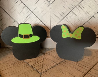 May include: Two black and green Mickey Mouse and Minnie Mouse heads with a green leprechaun hat on Mickey and a green bow with gold polka dots on Minnie.