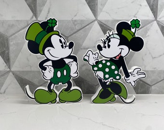 May include: A pair of cartoon figures, Mickey Mouse and Minnie Mouse, dressed in green and white outfits with shamrock accents. They are standing on a white surface.
