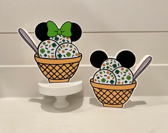 May include: Two wooden cutouts of Mickey Mouse's head with ice cream cones. The ice cream is white with green shamrocks and orange sprinkles. The cones are brown and have a waffle pattern. The Mickey Mouse heads are black with red bows.