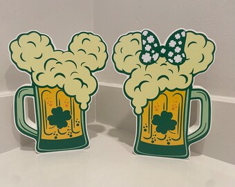 May include: Two green beer mugs with white foam and shamrock designs. One mug has a white bow with green polka dots on top.