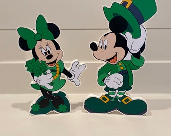 May include: Mickey Mouse and Minnie Mouse dressed in green leprechaun costumes. Mickey is wearing a green hat with a gold buckle and holding his hat in his hand. Minnie is holding a bouquet of green shamrocks.