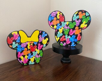 May include: A pair of wooden Mickey Mouse and Minnie Mouse head silhouettes. The silhouettes are decorated with a colorful pattern of balloons. The Minnie Mouse silhouette has a yellow bow.