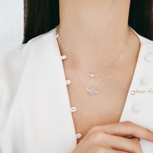 Puede incluir: Un collar plateado de dos capas con un pequeño colgante redondo y un colgante redondo más grande. El collar lo lleva una persona con una camisa blanca.