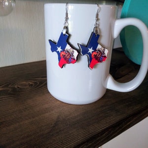 May include: A pair of earrings featuring a Texas state outline with a red, white, and blue design. The earrings are hanging from a white mug.