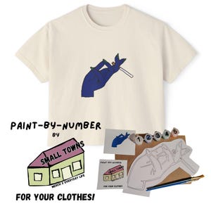 Puede incluir: Camiseta color crema con una ilustración de una mano azul sosteniendo un cigarrillo. La imagen incluye un kit de pintar por números con el texto "PAINT-BY-NUMBER BY SMALL TOWNS FOR YOUR CLOTHES!" y materiales de pintura.