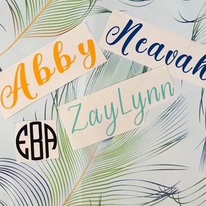May include: Four white vinyl stickers with names written in cursive. The names are Abby, Nevah, ZayLynn, and EBA. The stickers are on a background of green and white feathers.