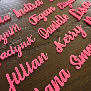 May include: Pink acrylic names, including Indra, Tegan, Danielle, Elizabeth, Lysa, Julian, Kerry, Alana, and  Sima, are displayed on a wooden surface.