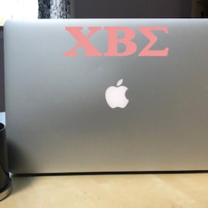 May include: A silver laptop computer with a pink Greek letter sticker that says "ΧΒΣ" on the lid. The laptop has a white Apple logo on the lid.