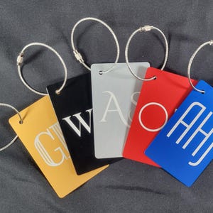 Könnte beinhalten: Fünf Gepäckanhänger mit Metallringen. Die Anhänger sind schwarz, gold, silber, rot und blau. Die Anhänger haben weiße Buchstaben, die "GW", "AS", "O", "AHJ" buchstabieren.