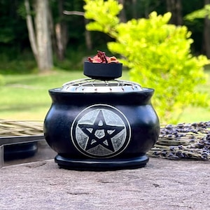 Peut inclure: Brûle-encens en pierre noire avec un motif de pentagramme. Le brûleur est doté d'une grille en métal et d'un petit bol sur le dessus pour l'encens. Des pétales de rose séchés sont dans le bol.