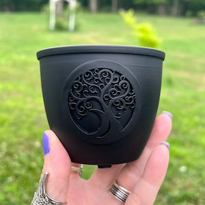 May include: A black, cylindrical container with a decorative tree of life design. The tree is embossed on a circular emblem on the side of the container. The container is held in a hand with purple nail polish and silver rings.