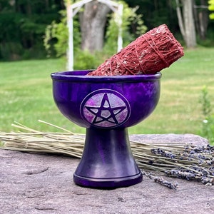 May include: A purple, pedestal-style bowl with a pentagram design holds a red sage bundle. The bowl sits on a stone surface with dried lavender and grass in the background. The bowl has a glossy finish.