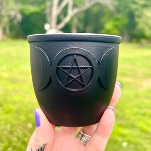 May include: A black ceramic cauldron-shaped cup with a white interior. The cup features a raised design of a pentagram within a circle, flanked by crescent moons. The cup is held against a blurred green background.