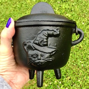 May include: A black cast iron cauldron with a lid and three legs. The cauldron features a raised relief of a cat wearing a witch's hat. The cauldron has a handle on the side and is held in a person's hand.