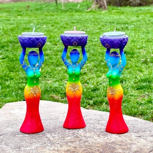 May include: Three tall candle holders shaped like human figures with a rainbow gradient finish from red at the base to blue at the top. Each figure holds a purple lotus-shaped bowl containing a tea light candle. Gold accents decorate the figures.
