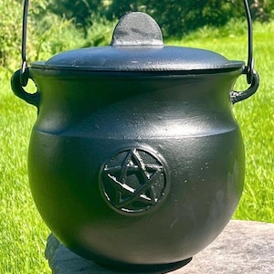 May include: A black cast iron cauldron with a rounded body, a matching lid with a small handle, and a metal swing handle attached to the sides. The front of the cauldron features a raised, circular pentagram symbol embossed on the surface.