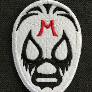 以下が含まれることがあります： 白と黒の刺繍パッチ。様式化されたマスクのデザインが特徴です。マスクには大きな目の開口部、口、中央に赤い「M」があります。パッチは楕円形で、衣類やアクセサリー用である可能性があります。