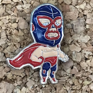 Puede incluir: Un pin de esmalte azul y rojo de un luchador con máscara y capa. El luchador está en una pose de lucha con un brazo levantado.