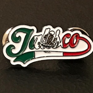 May include: A white enamel pin with the word "Taco" in green and red lettering. The pin features a stylized Mexican flag with the coat of arms in the center.