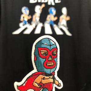 May include: A blue and red embroidered patch of a wrestler wearing a mask and cape. The wrestler is standing with his arms crossed and has a serious expression on his face. The patch is on a black background with the text "LIBRE" above it.