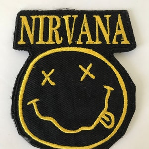 Puede incluir: Un parche bordado negro y amarillo con la palabra "NIRVANA" en letras amarillas sobre una cara sonriente amarilla con dos X negras para los ojos.