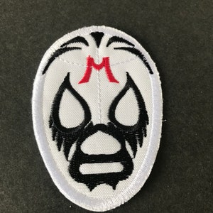Puede incluir: Parche blanco bordado con un diseño de máscara de lucha libre negra. La máscara tiene una "M" roja en la frente.