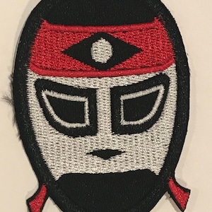 Puede incluir: Máscara de lucha libre bordada en blanco y negro con una cinta roja. La máscara tiene un ojo blanco y dos ojos negros.