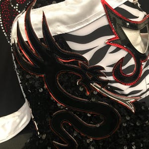 May include: Close-up of a black and red lucha libre mask with a dragon design. The mask features a black sequined base, a white and zebra-striped section, and red accents. The dragon is outlined in black with red detailing.