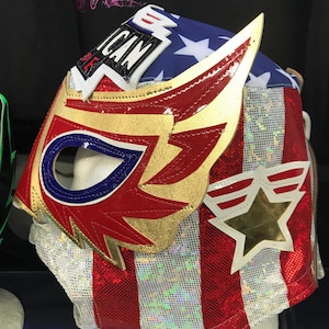 May include: A patriotic wrestling mask with a red, white, and blue color scheme. The mask features a star-spangled design, gold accents, and the word "AMERICAN". The mask is designed to be worn during wrestling matches.