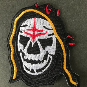 Peut inclure: Patch brodé noir avec un crâne blanc portant une robe à capuche rouge et jaune. Le crâne a une croix rouge sur le front.
