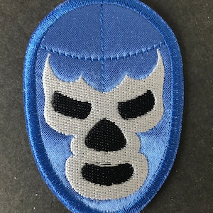 Puede incluir: Un parche bordado azul con un diseño de máscara de lucha libre estilizada. La máscara tiene una cara blanca y negra con un contorno azul. El parche es ovalado y tiene un borde azul, ideal para coser en ropa.