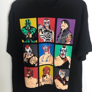 Pode incluir: Camiseta preta com uma grade de nove ilustrações coloridas de lutadores mascarados. Cada lutador é retratado com uma máscara e traje únicos, em um fundo de várias cores. A camiseta está pendurada em um cabide preto.