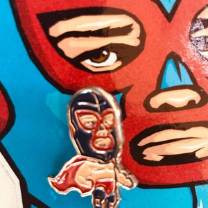 Puede incluir: Un pin de luchador de lucha libre azul y rojo con un contorno dorado. El luchador lleva una máscara azul con una franja roja y una capa roja. El pin es una ilustración de estilo de dibujos animados.