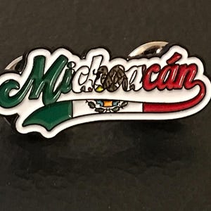 May include: A white, green, and red enamel pin with the word "Michoacán" in a stylized font. The pin features the Mexican flag with the coat of arms in the center.