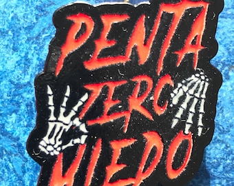 May include: A black and red enamel pin with the text "Penta Verc Miedo" in a stylized font. The text is outlined in red and the background is black. Two white skeleton hands are on either side of the text.