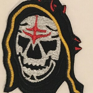 Puede incluir: Parche bordado negro con una calavera blanca que lleva una túnica negra con capucha y una cruz roja en la frente. La túnica tiene un borde amarillo y cuernos rojos.