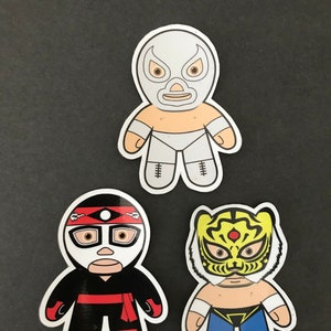 Puede incluir: Tres pegatinas de luchadores coloridos con contornos blancos. Las pegatinas presentan a un luchador enmascarado blanco, un luchador enmascarado negro con una cinta roja para la cabeza y un luchador enmascarado amarillo con un diseño de tigre.